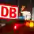El logo de DB se ve junto a un reloj y la indicación de parada de tranvía, mientras se atisban las luces de un vehículo.