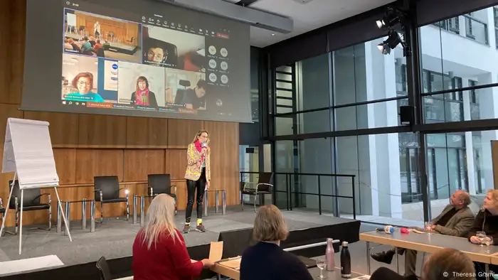  The woman on the stage, Zahra Nedjabat, speaks into a microphone in front of a group of listeners. On a projector screen behind her, people are taking part in the meeting via Teams video link.