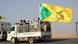 A white truck with a number of men standing on the bed at the back; one is holding a gun. A yellow and green flag with black writing flutters from the back of the truck.