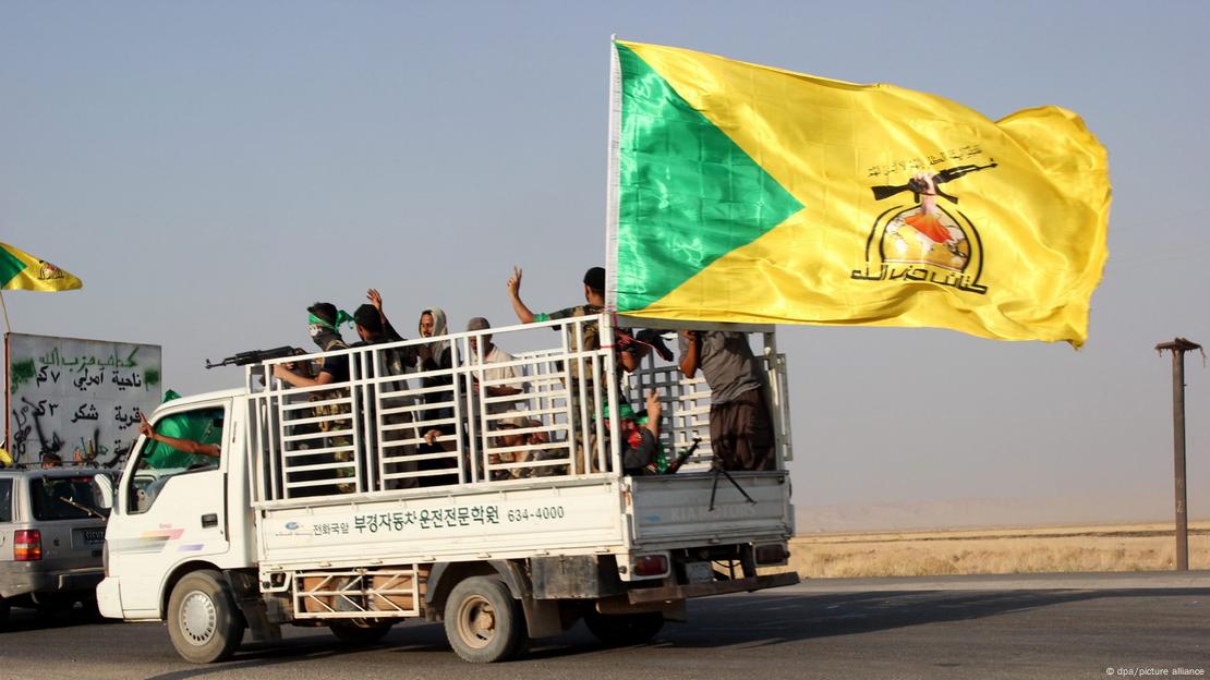 A white truck with a number of men standing on the bed at the back; one is holding a gun. A yellow and green flag with black writing flutters from the back of the truck.