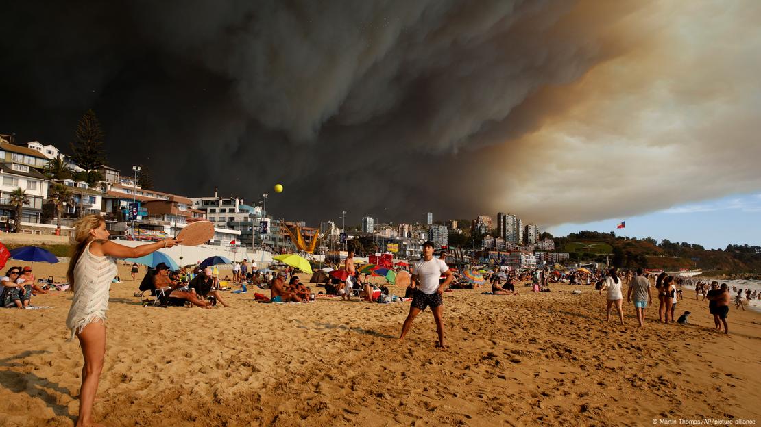 Duas pessoas jogam frescobol em praia, com o céu negro de fumaça