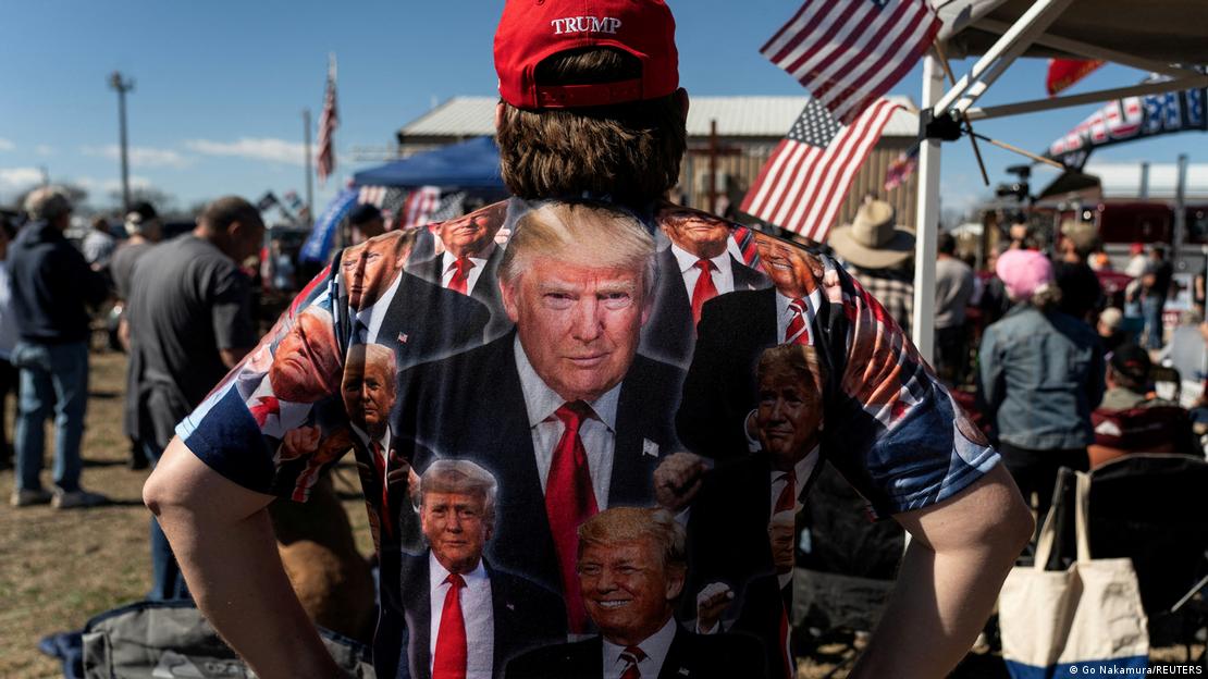 Homem de costas com camisa estampada com fotos de Donald Trump, em local de festa, com bandeiras dos EUA ao fundo