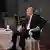 O apresentador de talk show americano, Tucker Carlson (esquerda), conduz uma entrevista com Vladimir Putin, presidente da Rússia. Ambos estão sentados em cadeiras brancas, frente à frente, com uma pequena mesa de centro entre eles.