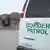 Im Vordergrund der geschlossene Kofferraum eines Autos mit der Aufschrift "Border Patrol" (Grenzpatrouille), dahinter steht eine Reihe Menschen