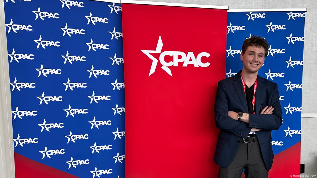 CPAC 2024: junger man steht vor CPAC-Banner und lächelt in Richtung Kamera