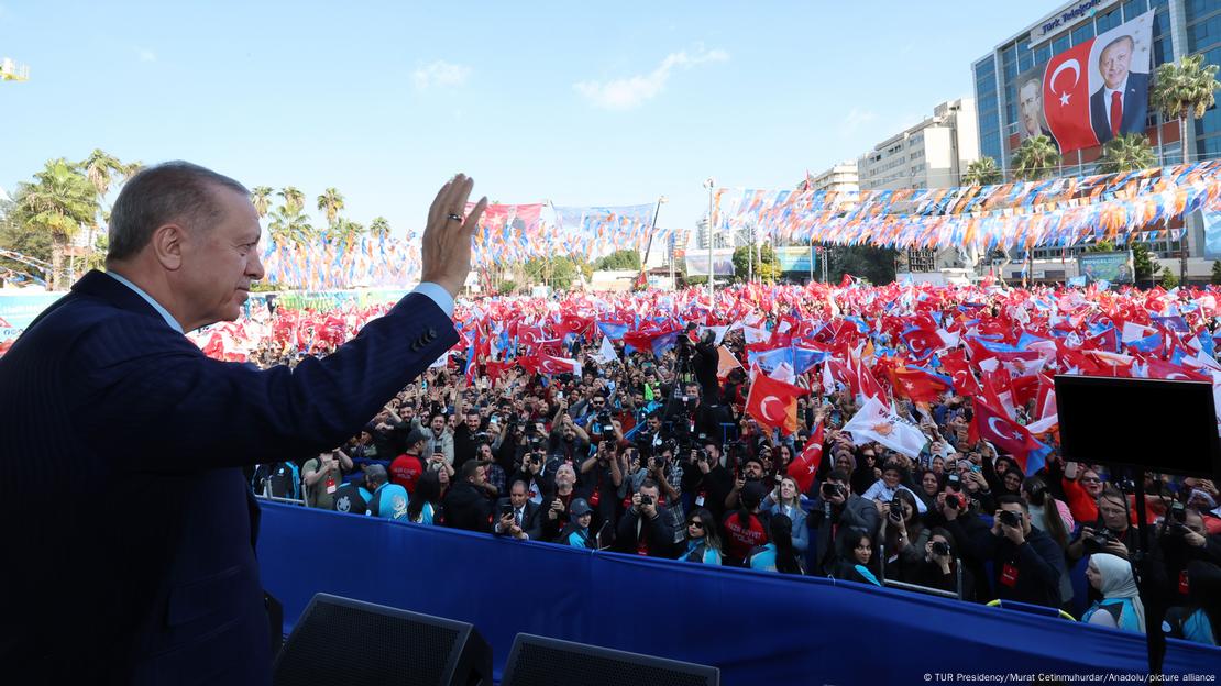 Erdogan steht in der südlichen Stadt Adana auf einer Bühne einer Wahlkampfveranstaltung. Vor ihm eine lebendige Menschenmenge mit vielen türkischen und AKP-Fahnen. Erdogan winkt mit dem rechten Arm zum Abschied.