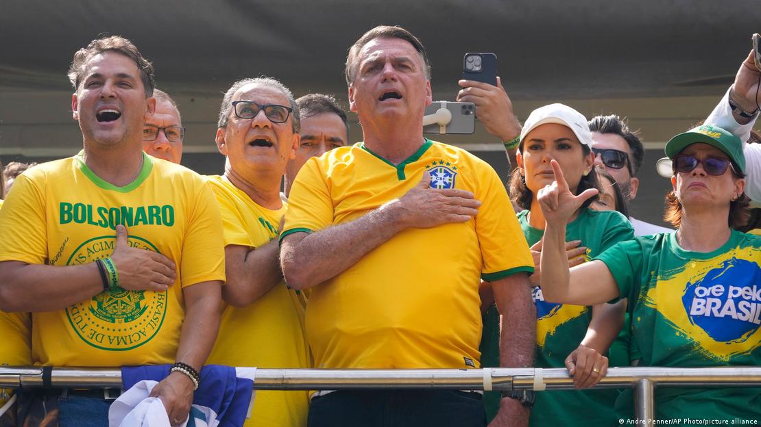 Bolsonaro com camiseta da seleção brasileira de futebol canta com a mão no coração, ao lado da esposa e outros apoiadores