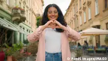 Smiling happy young teen Indian Arabian woman female lady girl making heart shape hands to camera laughing positive gesture love like emoji support care sign symbol standing outside town city street. High quality 4k footage