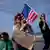 Three women — one holding a cellphone, one an American flag, and another a child and a sign reading 'End This Deadly War' — are seen protesting at a beach in Tel Aviv in March 2024.