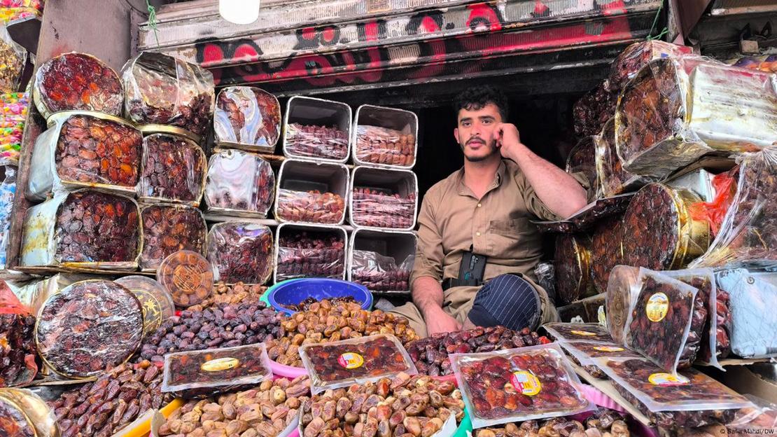 A vendor is sitting in his stall at the market, nuts and dates in front of him