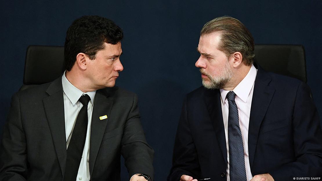 Sergio Moro e Dias Toffoli sentados e se olhando, com um fundo preto