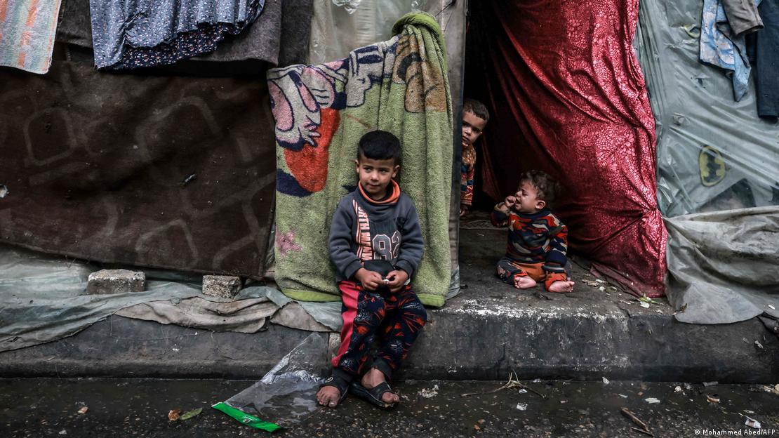 Três crianças na entrada de um abrigo improvisado com lonas e cobertores