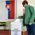 Una mujer deposita su voto en una urna mientras se aprecia una bandera eslovaca en la pared.