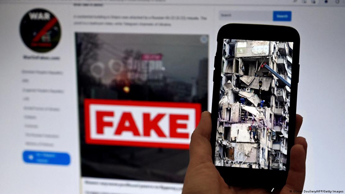 Tela de computador mostra a palavra "fake" sobre uma foto. Em primeiro plano, a foto de um prédio destruído é exibida em um smartphone