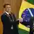 Emmanuel Macron e Luiz Inácio Lula da Silva dão as mãos em frente à bandeira do Brasil