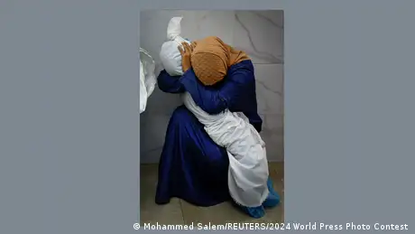 Eine Frau hält einen in weißen Stoff gewickelten Leichnam auf ihrem Schoß. Ihr eigener Kopf ist von einem Tuch verhüllt. 