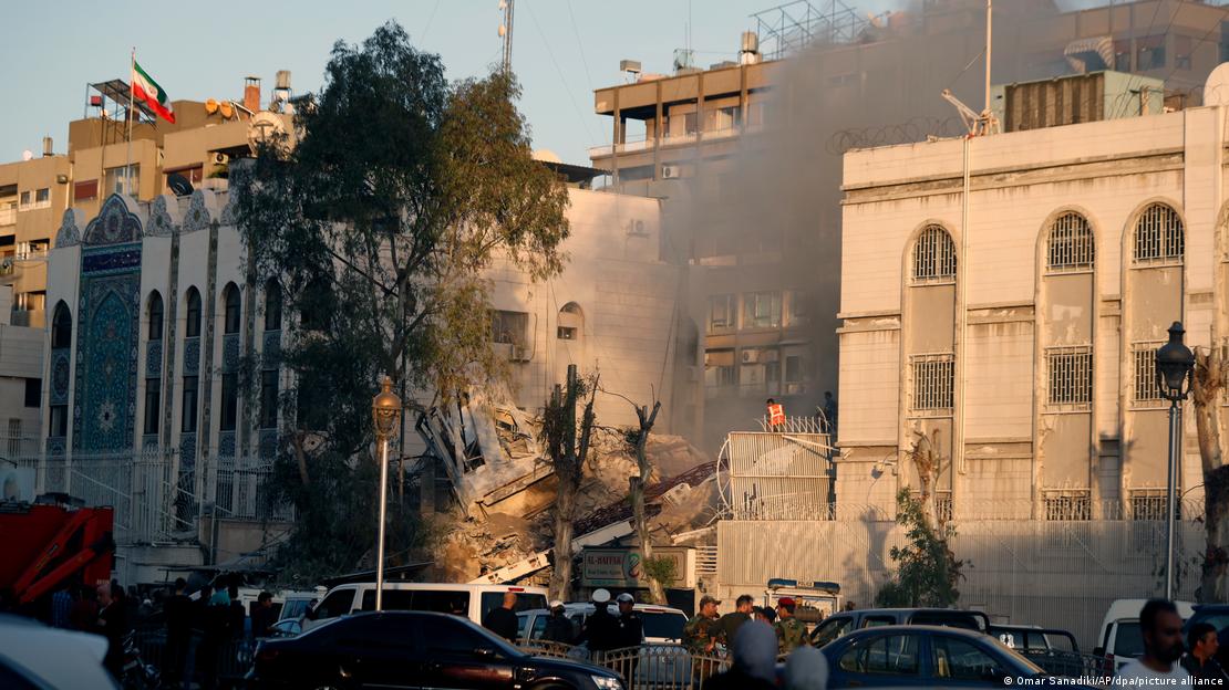 Escombros de um prédio com fumaça sobre ele, ao lado de outros edifícios intactos