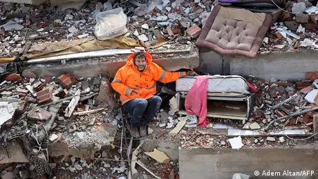 Ein Mann sitzt in einem neonorangenen Anorak zwischen Trümmern und hält die Hand einer Person, die unter Trümmern liegt. 