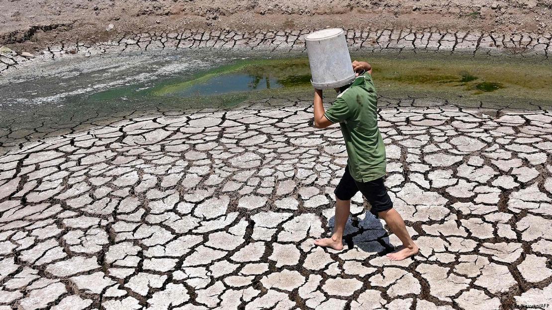 Başına plastik bir kova geçirerek kendini güneşten koruyan biri, çatlamış toprak zeminin üstünde yürüyor