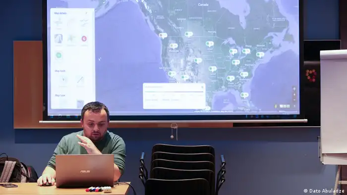 A man sits at a desk on his laptop. Behind him on a screen is a map with dot points located on it. 