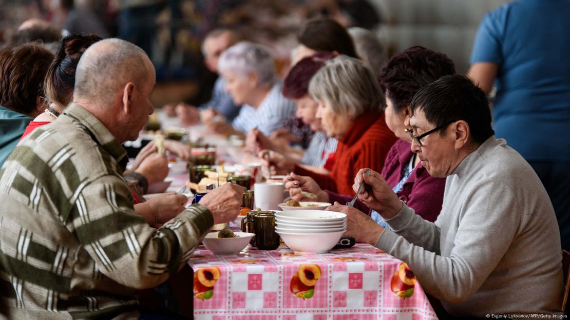 Evacuated people are sitting at a big table and having meals in their temporary housing. 