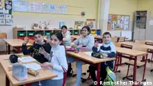 Blick in ein Klassenzimmer, in dem fünf Kinder an ihren Pulten sitzen und lächelnd in die Kamera schauen