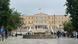 Der Syntagma-Platz im Athener Stadtzentrum mit dem Parlament im Hintergrund. Auf dem Dach des Parlaments weht die griechische Fahne. Auf dem Platz laufen Menschen. Der Syntagma-Platz im Athener Stadtzentrum mit dem Parlament im Hintergrund. Auf dem Dach des Parlaments weht die griechische Fahne. Auf dem Platz laufen Menschen.