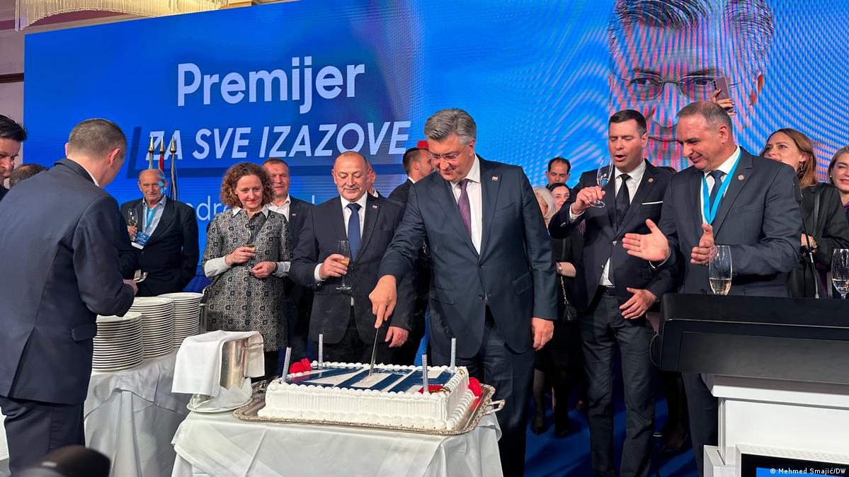 Ein Mann in Anzug steht in einer Gruppe formell gekleideter Menschen und schneidet einen Kuchen mit den Buchstaben HDZ an.