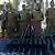 About a dozen soldiers in plain olive green uniforms stand to attention in a row, facing the camera, with several rifles in front of them