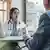 Boynunda stetoskop asılı bir kadın doktor, sırtı dönük bir kişi ile masa başında konuşuyor