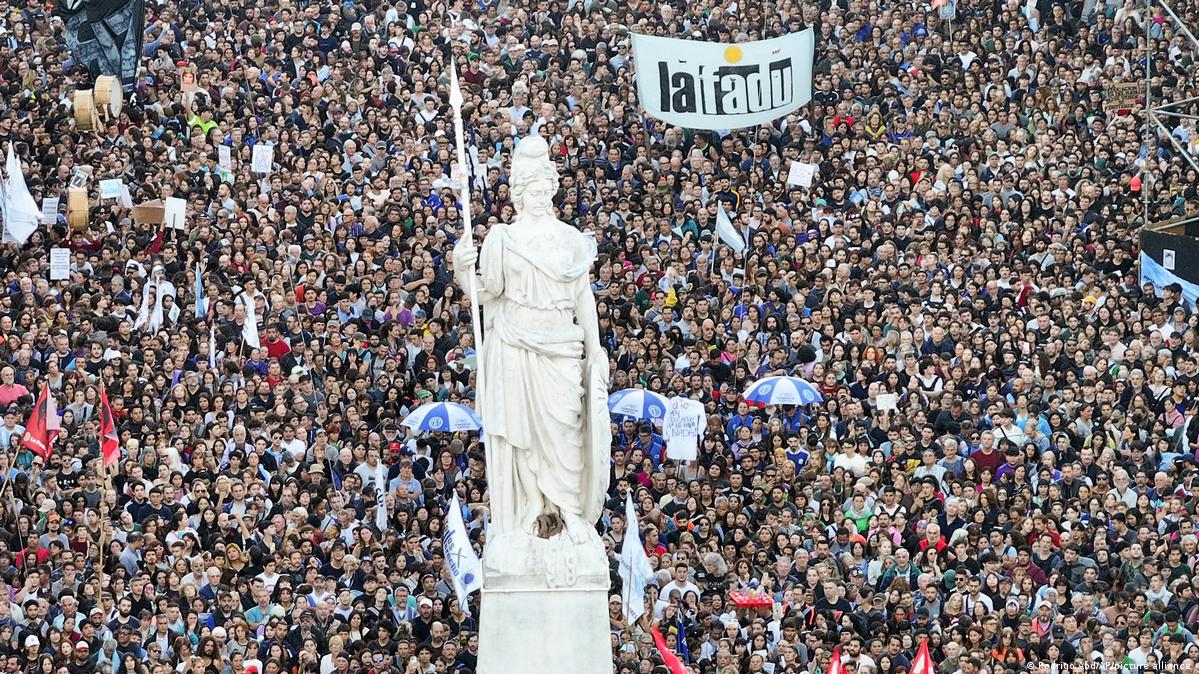 A large crowd of people stand together during a demonstration behind a giant statue