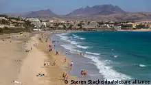 Urlauber gehen am Strand der Costa Calma am Meer spazieren. Etwas oberhalb des Strandes reihen sich die Hotels aneinander.