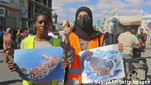 Two women in brightly colored safety vests, one holding a photo and the other a painting, both depicting migrants in boats at sea
