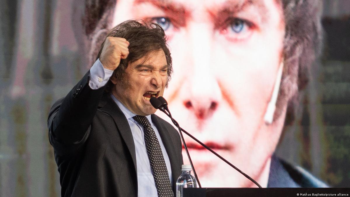 Argentine President Javier Milei raises his fist as he yells into a microphone in front of a large screen with his likeness upon it 
