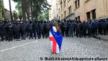 14.05.2024, Georgien, Tiflis: Ein Demonstrant mit drapierten georgischen National- und EU-Flaggen steht vor der Polizei, die den Weg zum Parlamentsgebäude blockiert, während eines Protestes der Opposition gegen das «russische Gesetz». Begleitet von Protesten hat das georgische Parlament die Verabschiedung eines umstrittenen Gesetzes begonnen, das ausländischen Einfluss auf die Zivilgesellschaft beschneiden soll. Foto: Shakh Aivazov/AP/dpa +++ dpa-Bildfunk +++