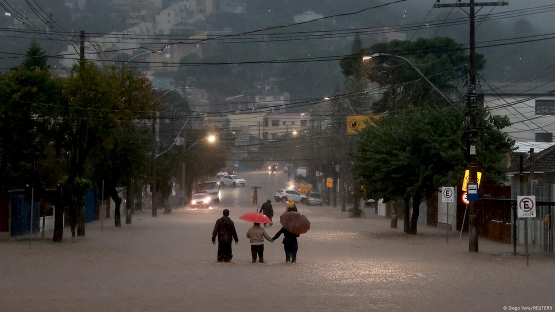 Imagem de pessoas caminhando numa rua coberta por água, nas enchentes que afetaram o Rio Grande do Sul em maio