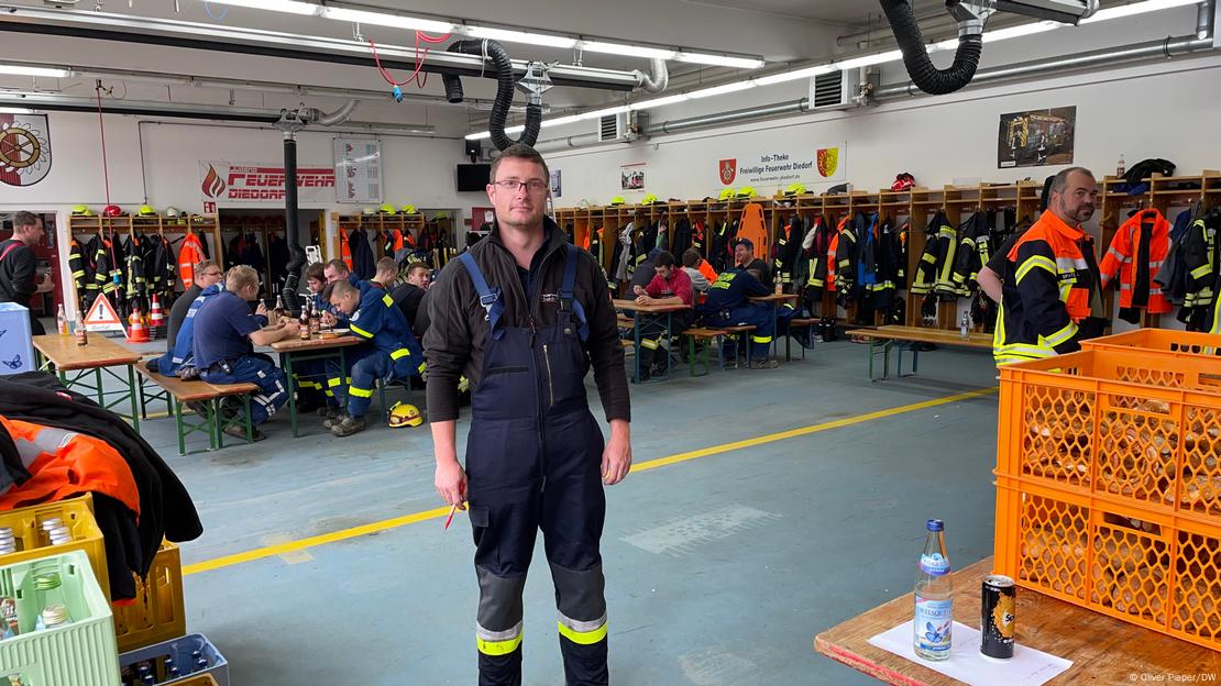 Mann steht in großer Halle im Blaumann, im Hintergrund Feuerwehrleute