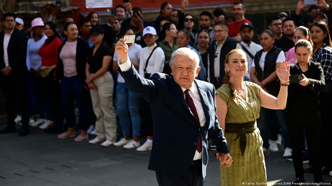 A well-dressed man and woman (Mexican President Andres Manuel Lopez Obrador and his wife Beatriz) wave and show a voting card as they walk past a crowd of onlookers