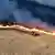 Luftaufnahme eines Feldes, dessen hinterer Teil in Flammen steht - vor der Feuerwand fährt ein Bulldozer durchs verdorrte Feld und schlägt eine Brandschneise