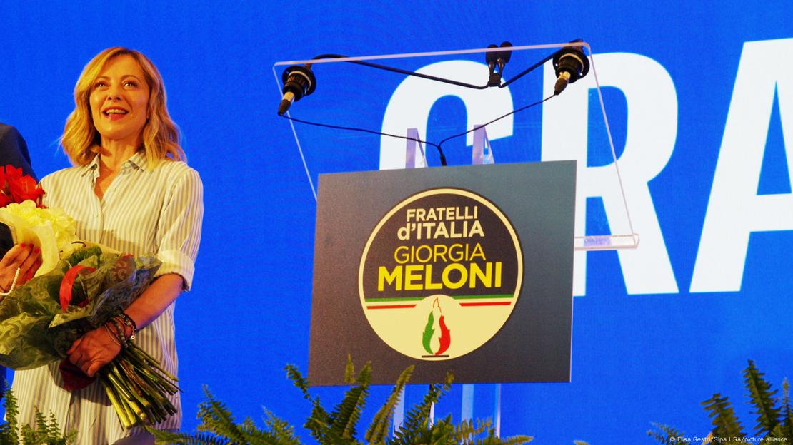 Giorgia Meloni steht mit einem Blumenstrauß neben einem Rednerpult mit dem Logo ihrer Partei und ihrem Namen darauf