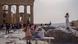 A tourist uses a hand fan to cool down another one sitting on a bench in front of the Parthenon at the ancient Acropolis, in Athens