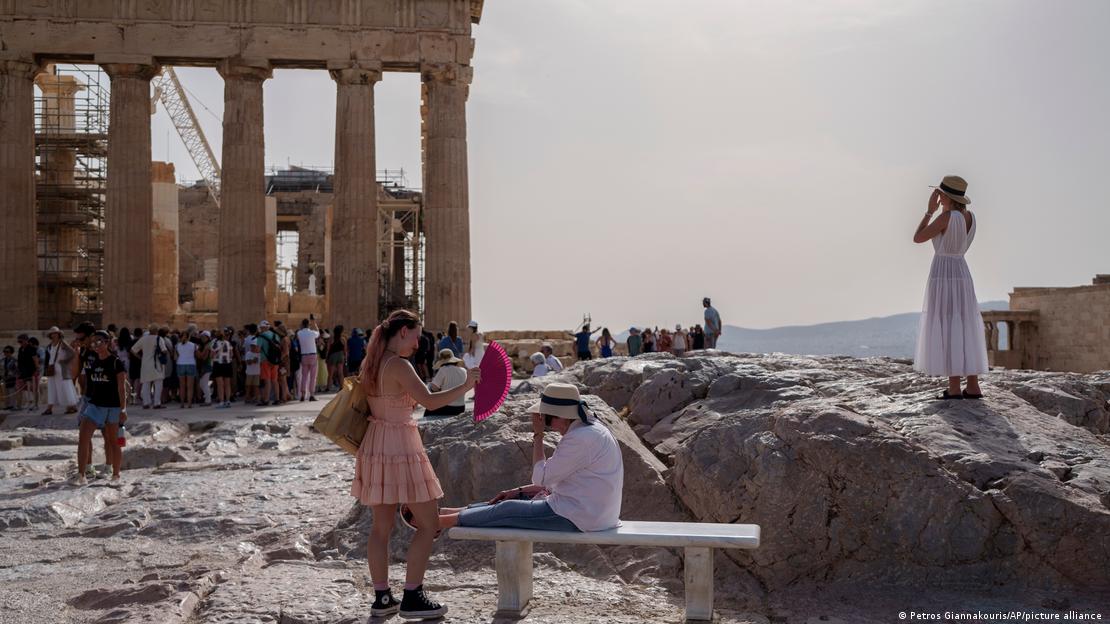 A tourist uses a hand fan to cool down another one sitting on a bench in front of the Parthenon at the ancient Acropolis, in Athens