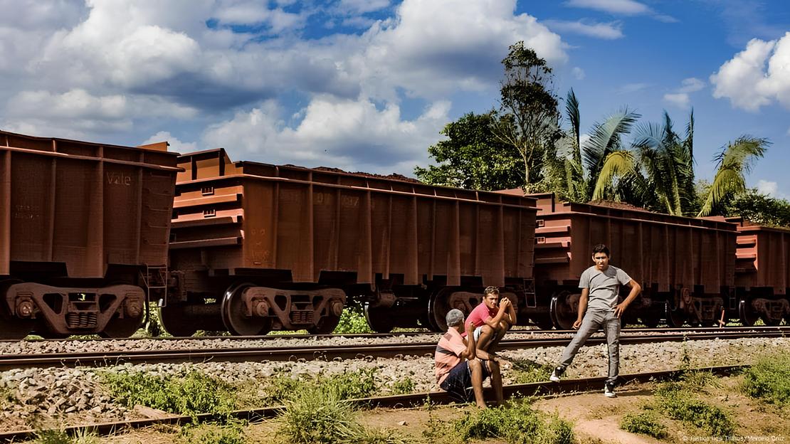 Três homens jovens sentados nos trilhos da Estrada de Ferro Carajás, com vagões de minério ao fundo, céu azul e algumas árvores espaçadas. Três homens jovens sentados nos trilhos da Estrada de Ferro Carajás, com vagões de minério ao fundo, céu azul e algumas árvores espaçadas.