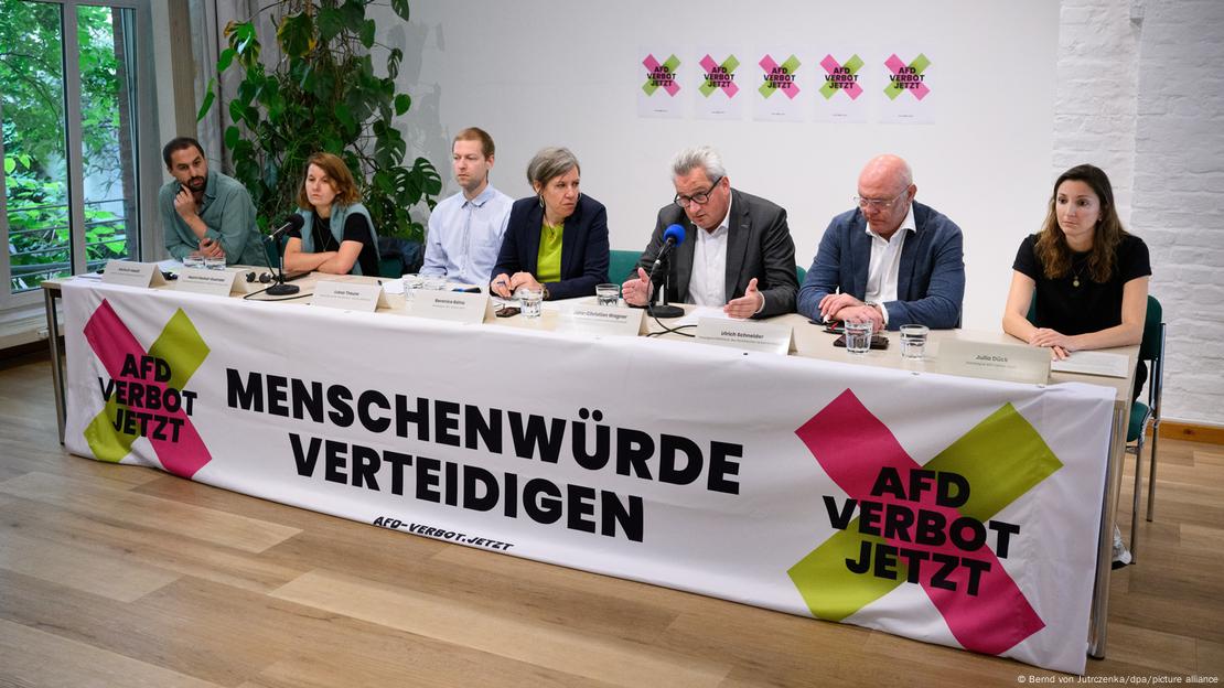 Sieben Personen sitzen an einem langen Tisch mit der Aufschrift "Menschenwürde verteidigen"