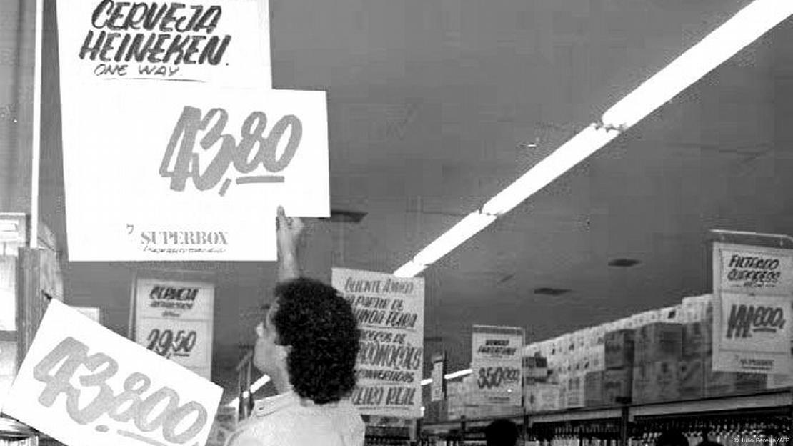 Foto preto e branco mostra homem de costas entre gôndolas de supermercado trocando placas de preço