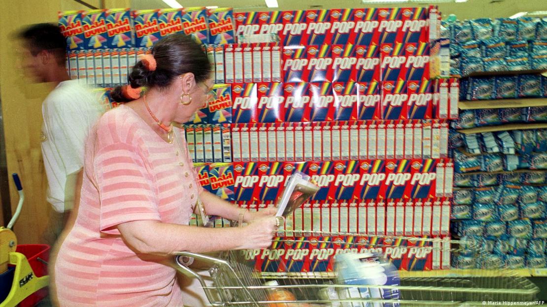 Mulher empurra carrinho de compras enquanto olha produtos de limpeza dispostos em uma gôndola