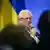Presidente del fabricante de armas alemán Rheinmetall, Armin Papperger, habla a un micrófono, con la bandera de Ucrania al fondo, y siluetas de varias personas onservándolo en primer plano.