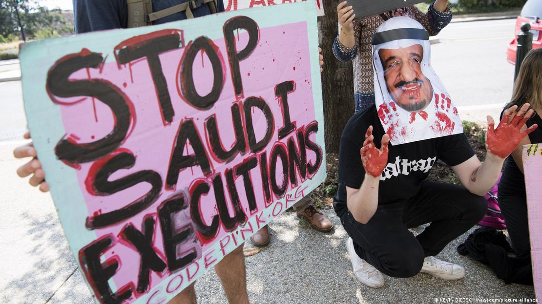 Pessoa segura cartaz com os dizeres "Pare as execuções sauditas” ao lado de outro manifestante com uma máscara de Al Saud e as mãos pintadas de vermelho.