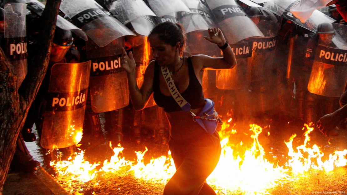 Barreira policial diante do rastro de fogo deixado por um coquetel molotov e, em primeiro plano, uma mulher que foge das chamas