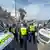 Police face off with a crowd of people on the shoreline, framed by two police vehicles